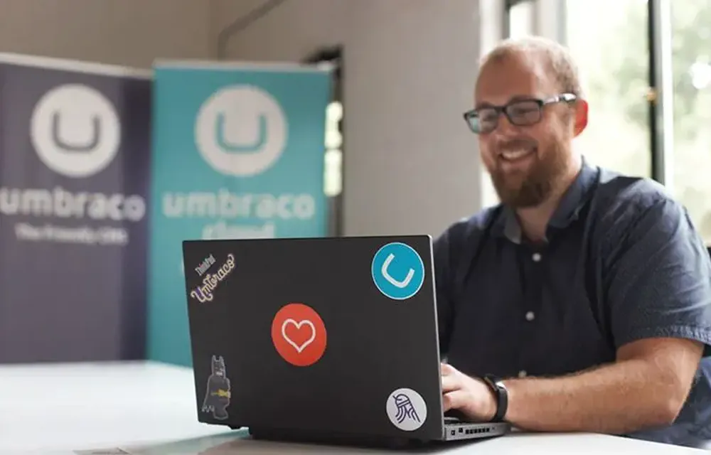 A man working on a laptop with Umbraco stickers on it, with Umbraco rollup banners in the background