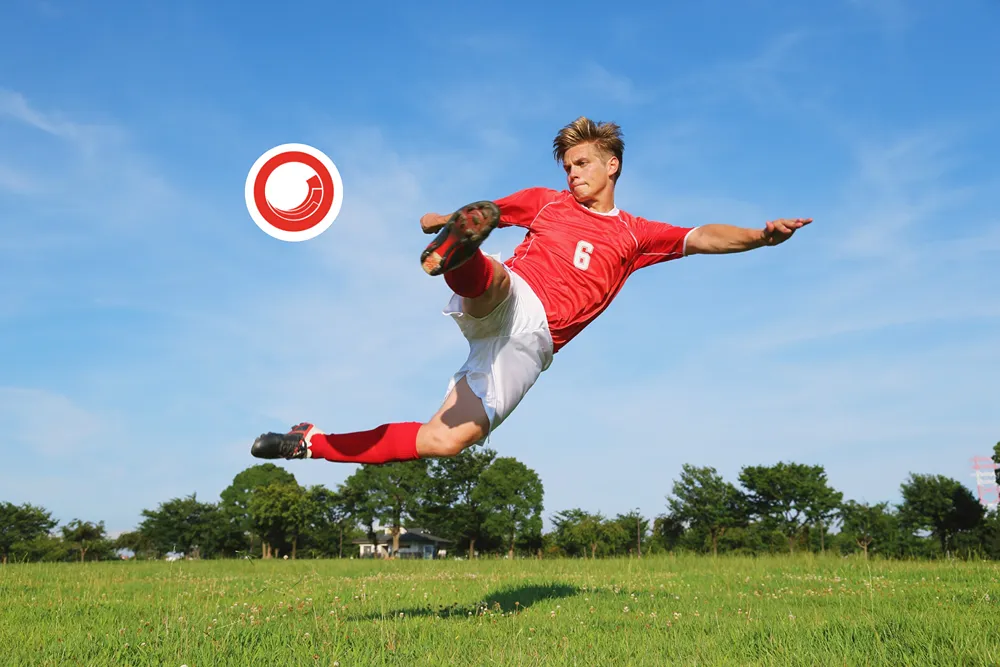 A footballer wearing a red football shirt and white shorts cross kicking a Sitecore football to score a goal. The background features a green field and low treeline.