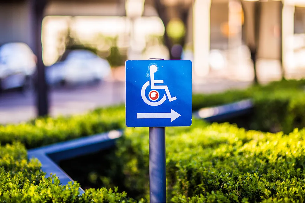 A disabled parking spot sign in a green hedge, with a Sitecore logo superimposed in the wheel section of the wheelchair glyph.