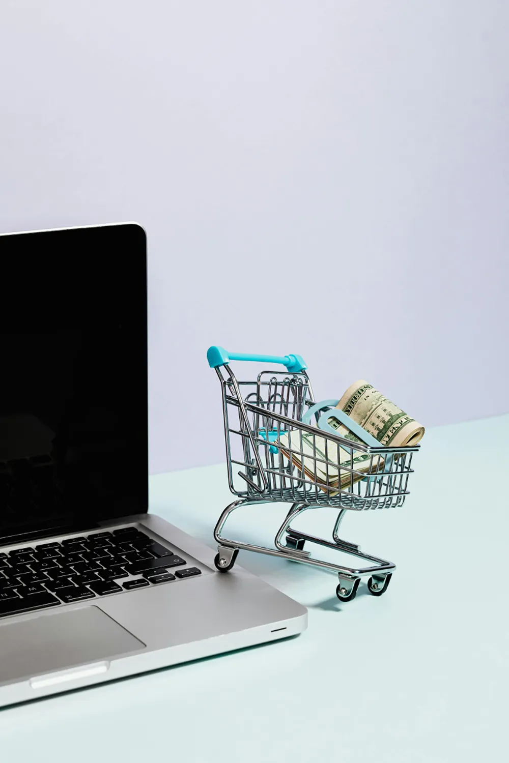 A small toy shopping cart full of American Dollar bills to the right of a laptop