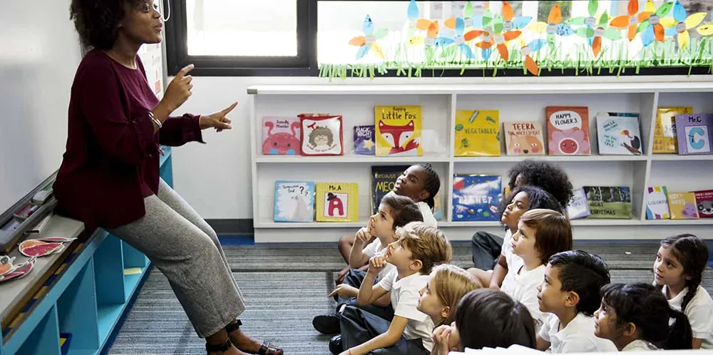 A teacher leading a class of young children, with a small library of books in the background.