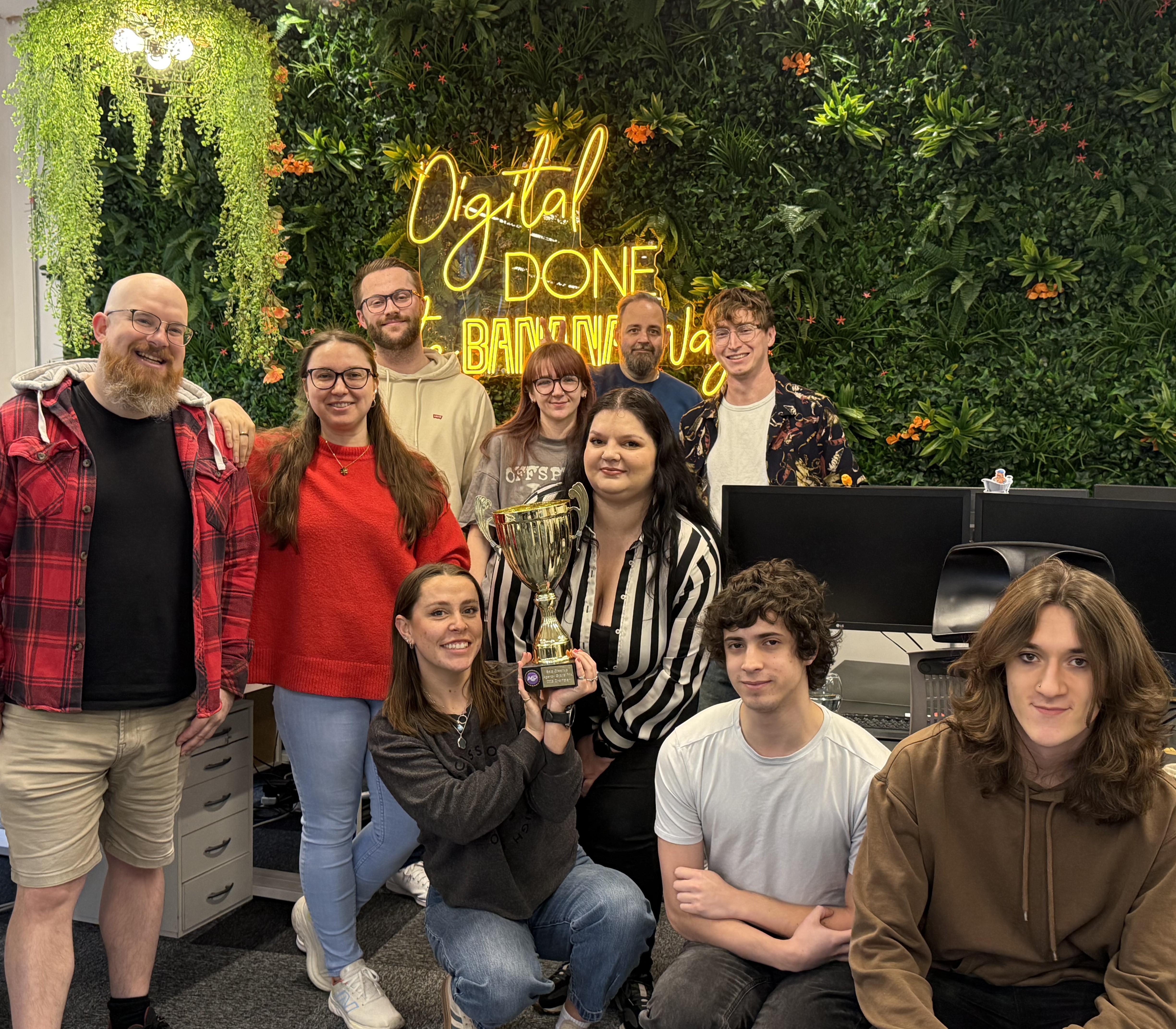 The Cold Banana Bournemouth HQ office team holding the Agency Grand Prix 2025 trophy in front of a wall with greenery and a yellow neon sign reading "Digital Done the Banana Way"