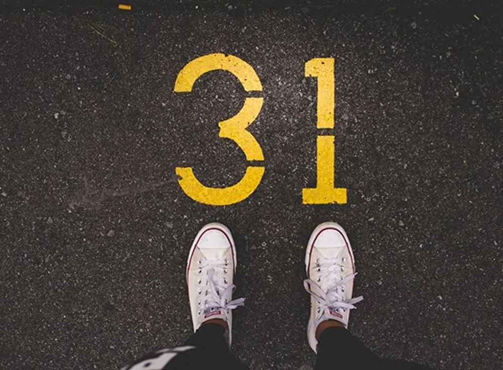 A person looking down at their white shoes with the number 31 written on tarmac in yellow paint.