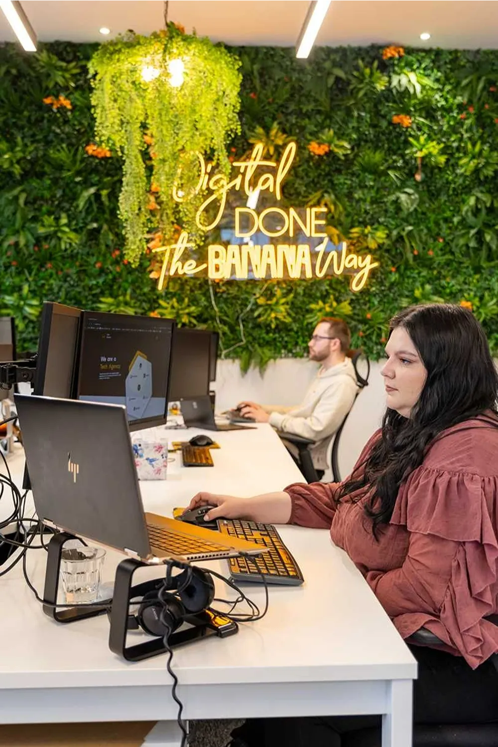 Cold Banana employees working at their desks with the Cold Banana green wall in the background, with a yellow neon sign reading "Digital Done the Banana Way"
