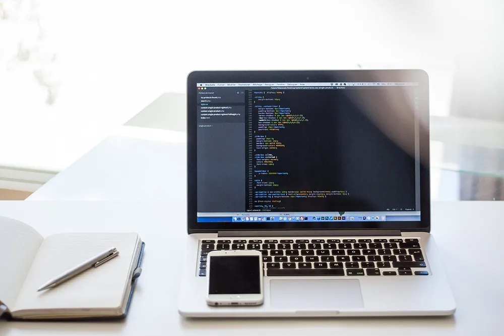 Code on a laptop, with a white smartphone on the keypad, and a notepad & pen to the left.