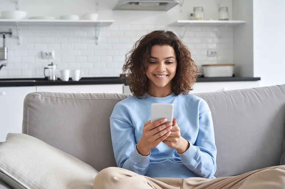 A person wearing a blue cardigan and sitting on a grey sofa looking at their smartphone.