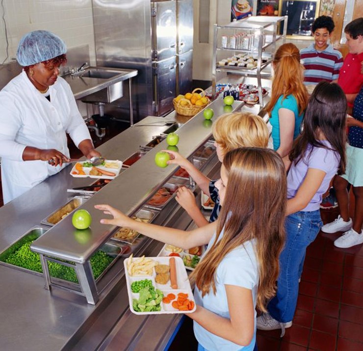 A school lunch line with a lunch lady serving the children food. In the foreground is a girl with a plate of food reaching for a green apple.