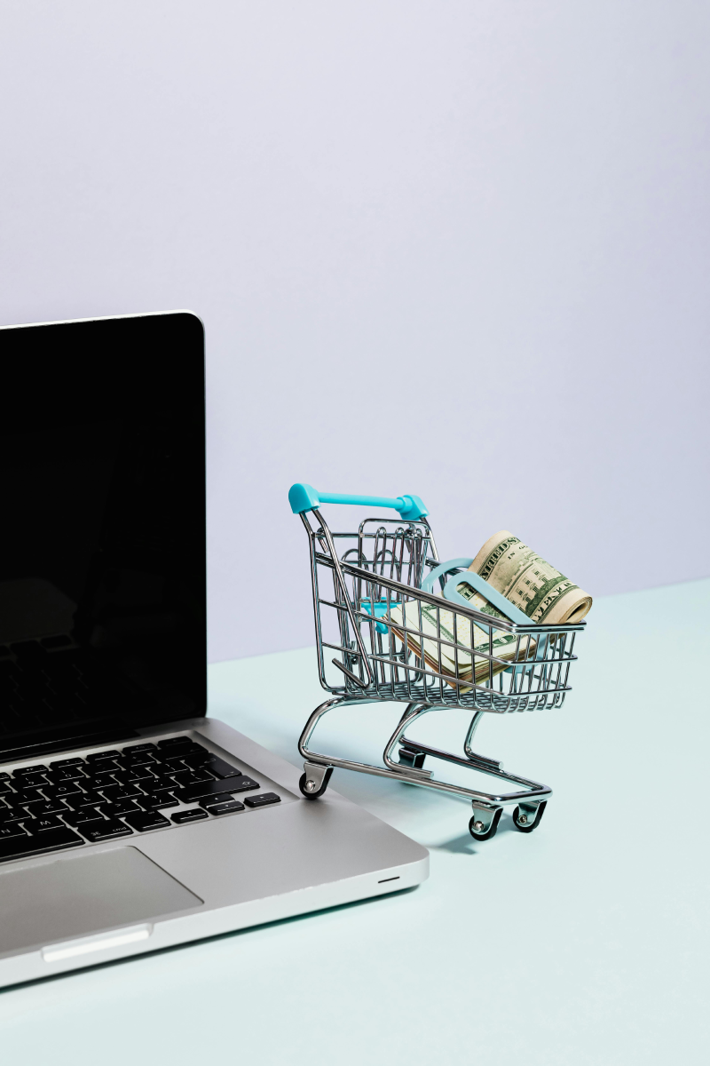 A mini toy shopping cart filled with a clip of American money perched on the edge of a silver laptop.
