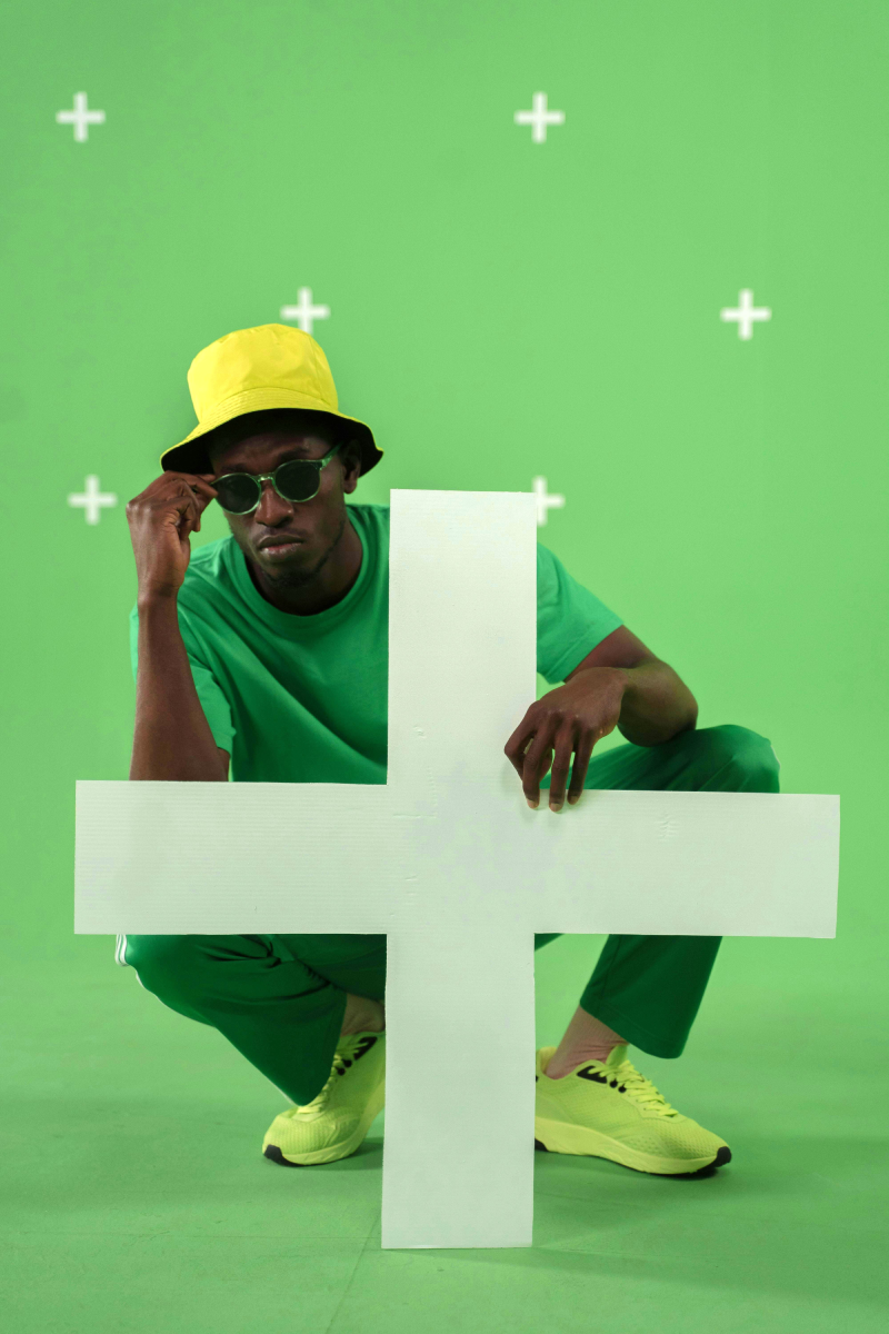 A man dressed in green crouching whilst holding a large white plus cutout, in a green room with plus signs on the walls