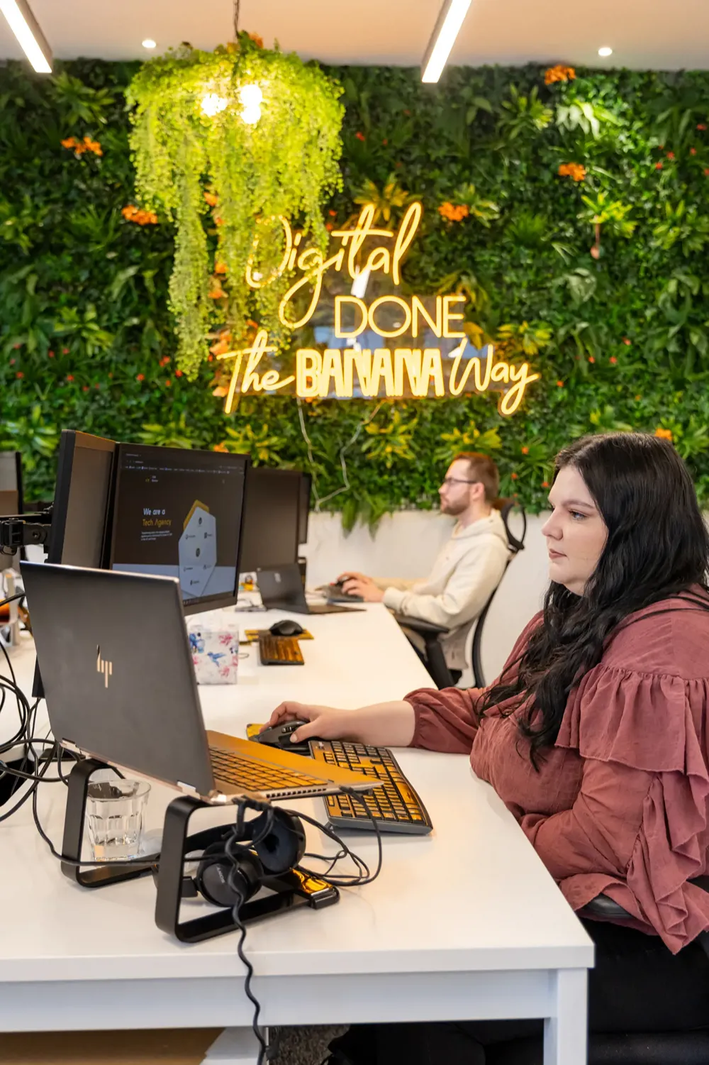 Cold Banana employees working at their desks with the Cold Banana green wall in the background, with a yellow neon sign reading "Digital Done the Banana Way"