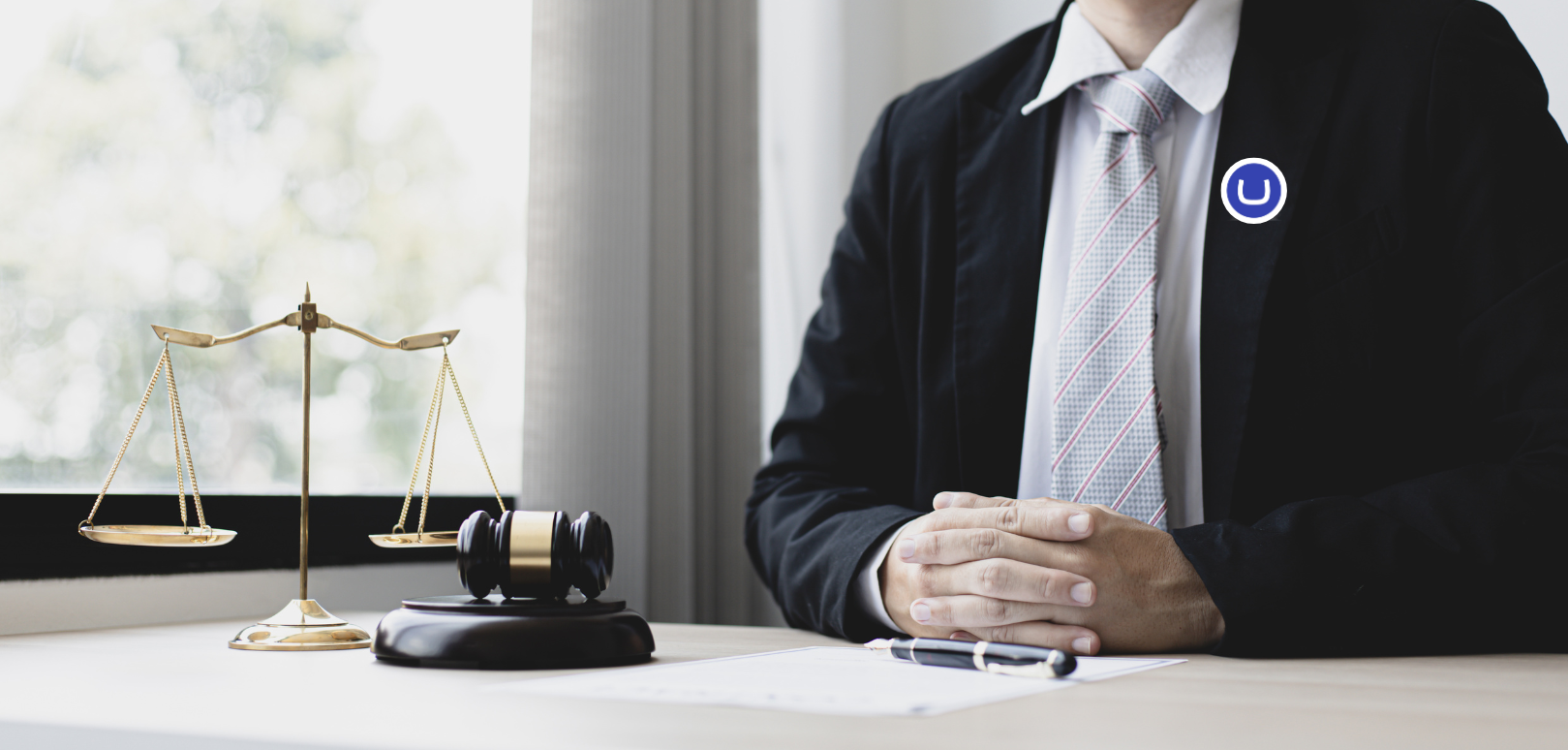 A lawyer in a suit sits behind a light brown desk with a superimposed Umbraco badge. In the foreground is a piece of paperwork, a pen, a gavel and a set of gold scales.