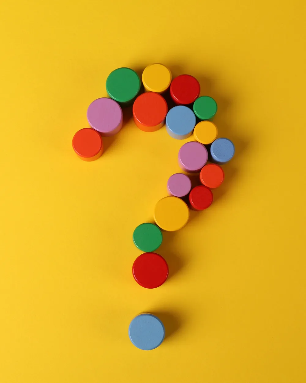 A question mark comprised of multicoloured wooden cylinders, on a yellow background.